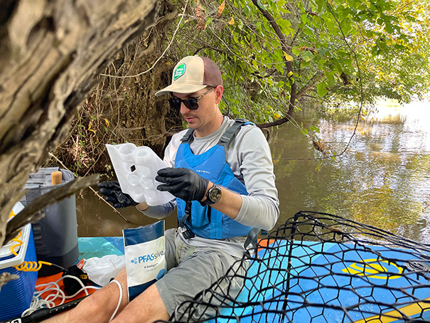 Thomas Dunlap sits on a raft next to the riverbank, holding a water sample container